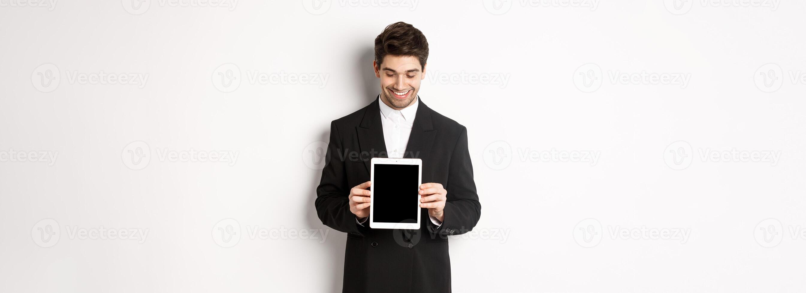 Image of good-looking male entrepreneur in black suit, looking down at digital tablet screen and showing advertisement, standing against white background photo