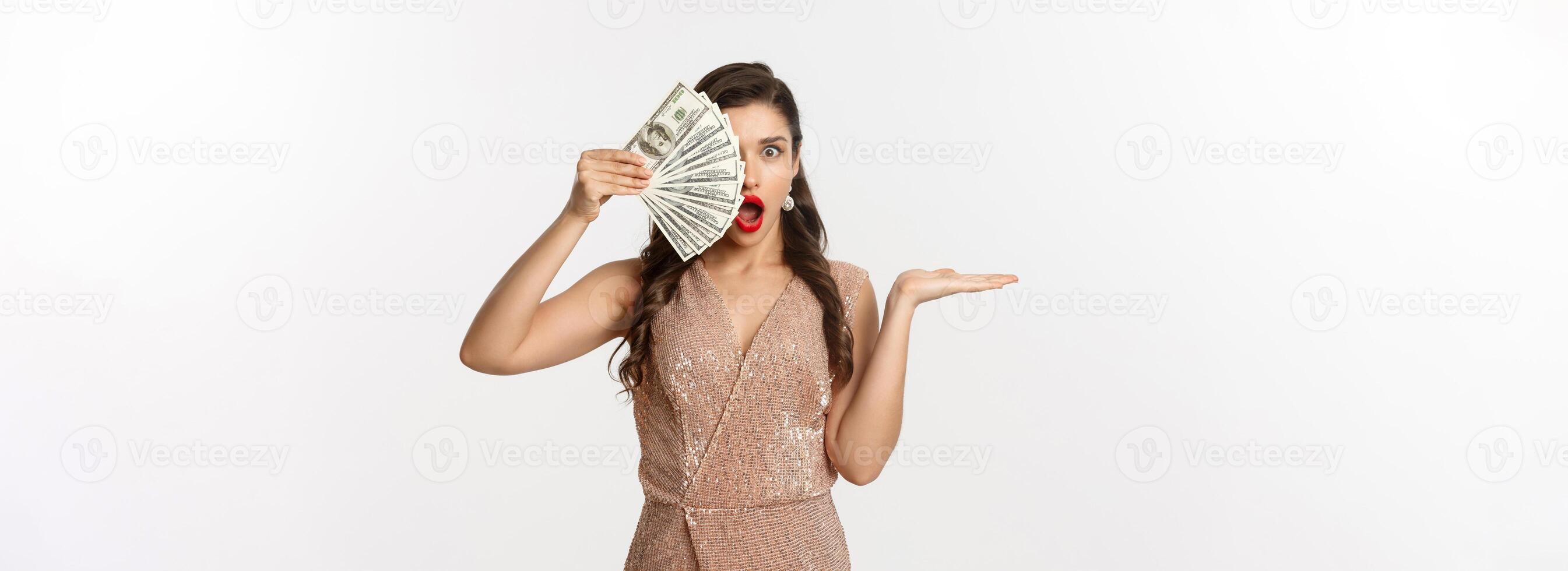 Shopping concept. Shocked woman holding money near face, wearing stylish dress, showing dollars with amazed expression, standing over white background photo