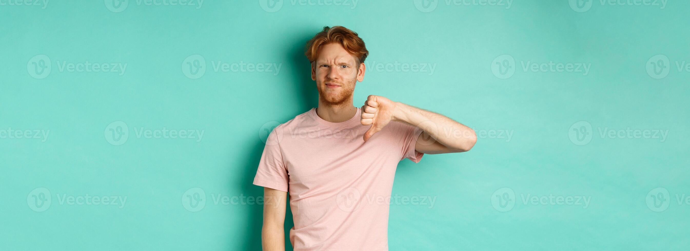 Disappointed redhead man showing thumbs-down, frowning and looking skeptical, epxress dislike and disapproval, standing over turquoise background photo