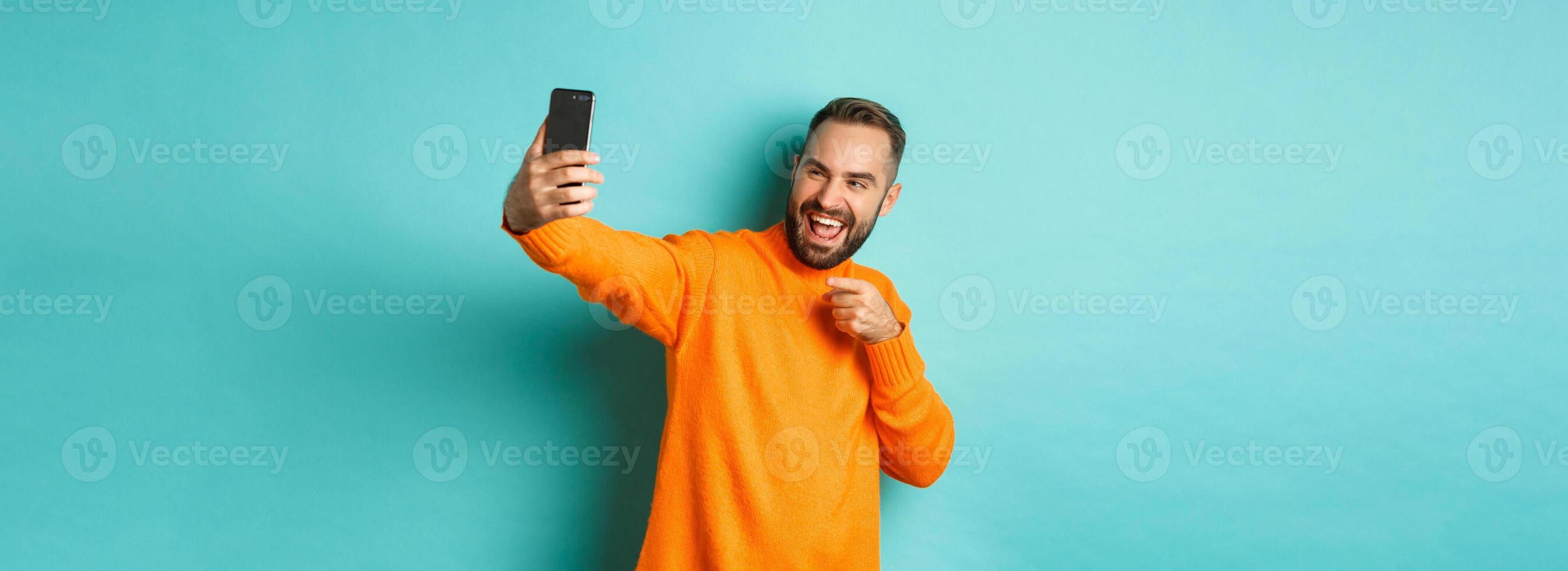 Handsome man calling on mobile phone, taking selfie and pointing at smartphone camera, standing over turquoise background photo