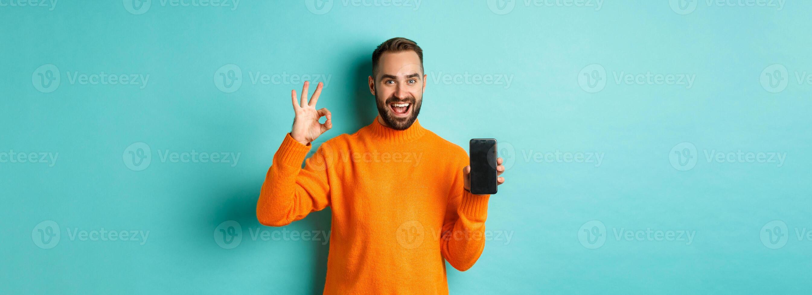 Photo of caucasian man showing mobile screen and okay sign, approve online store, smartphone app, standing satisfied over light blue background