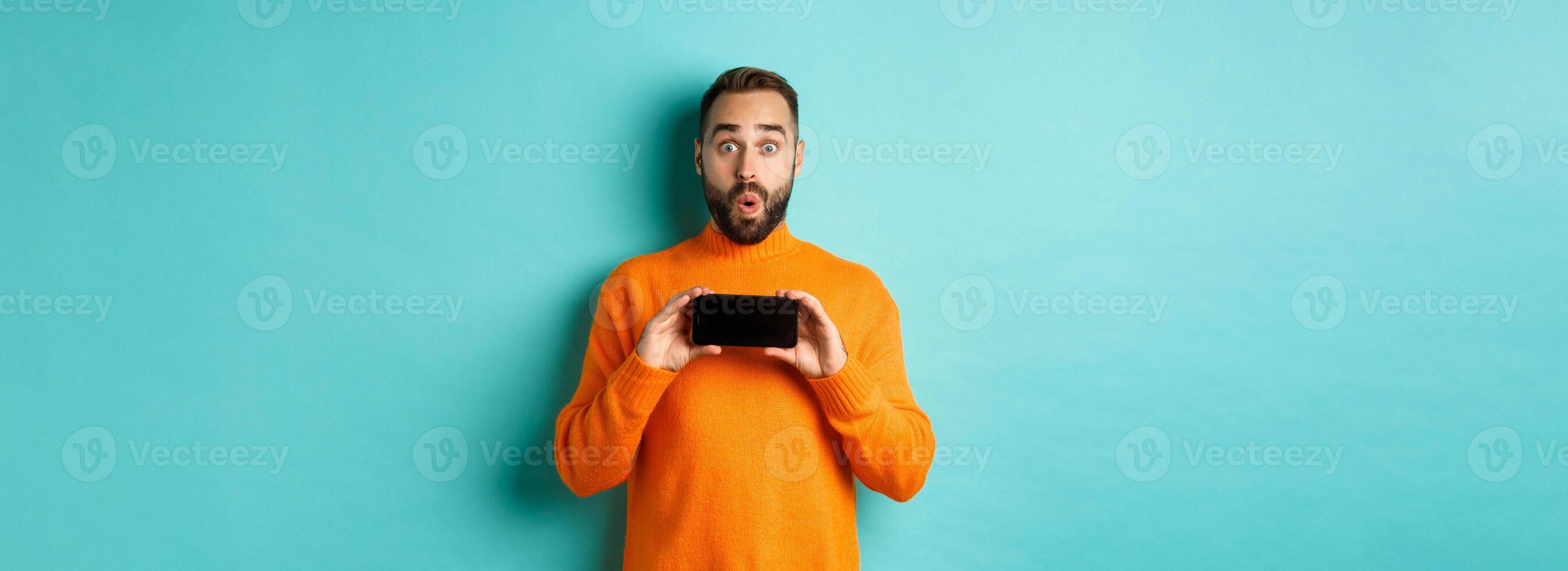 Impressed man showing smartphone screen, stare at camera amazed, demonstrate display, standing over light blue background photo