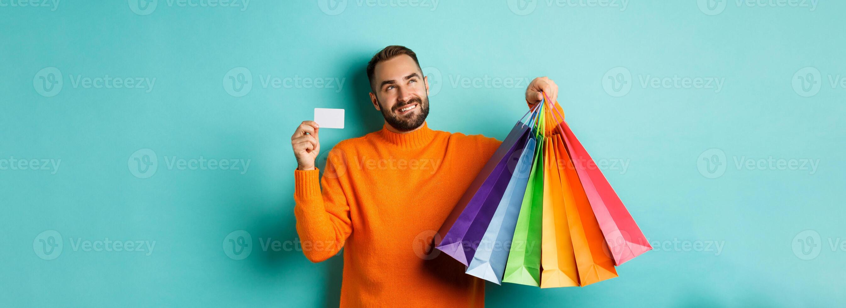 Happy aduly man showing credit card and shopping bags, standing against turquoise background photo