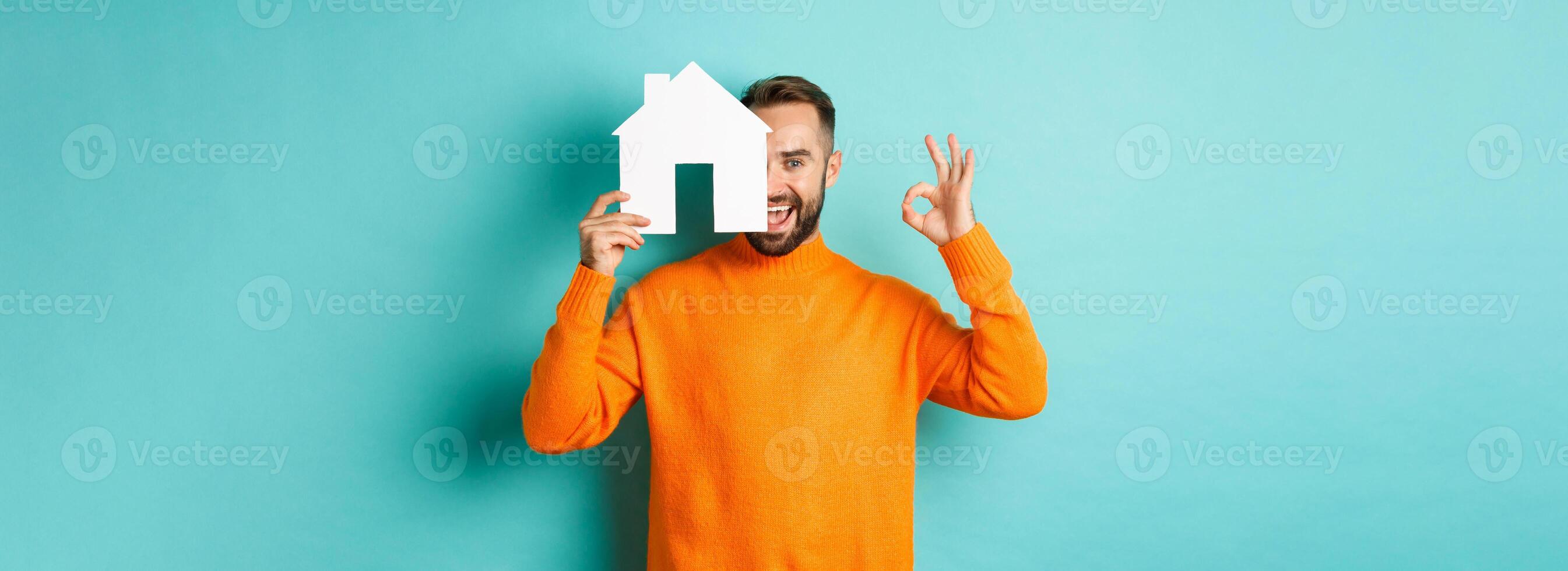 Real estate concept. Satisfied man recommending agency, showing okay sign and house maket, standing over blue background photo