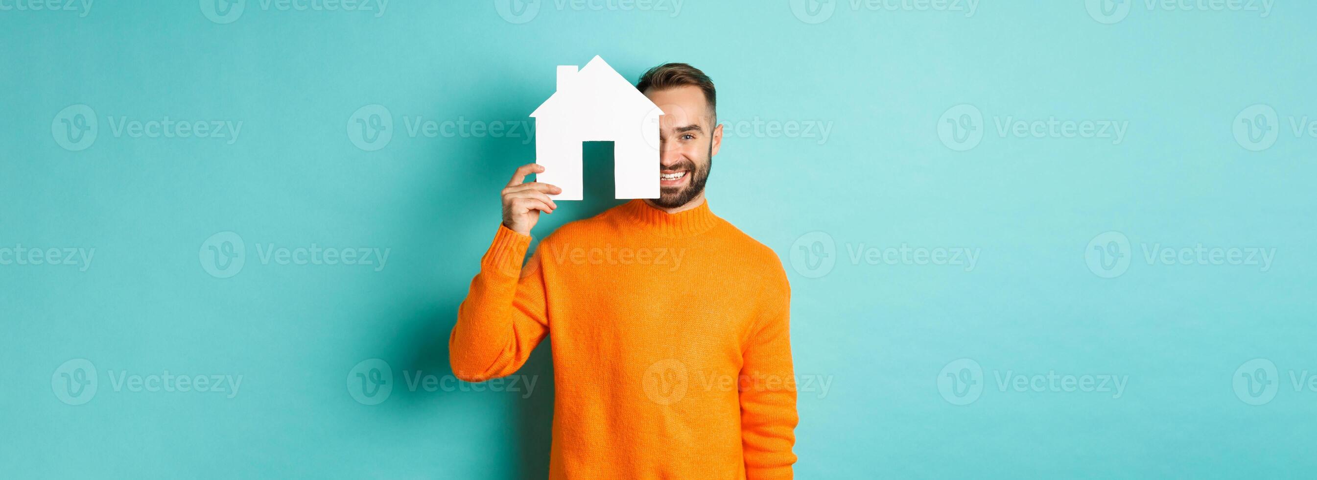 Real estate concept. Happy young man searching for home rent, holding house paper maket and smiling, standing over blue background photo