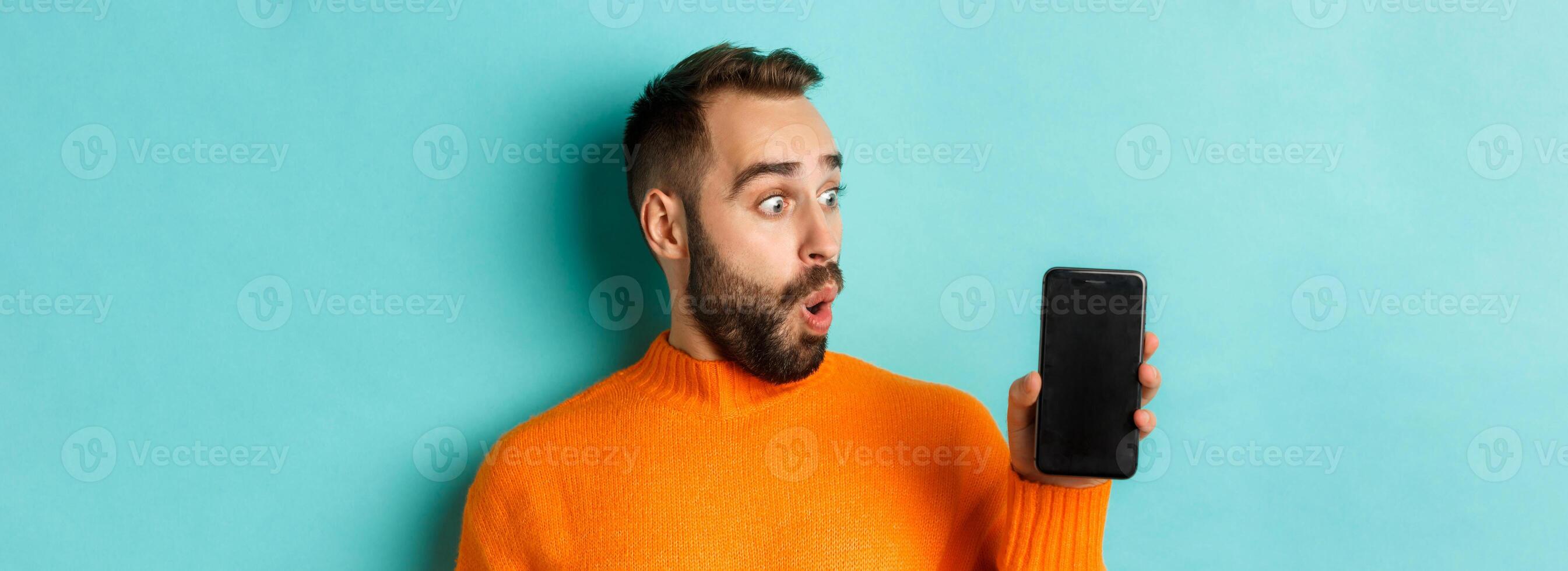 Close-up of young bearded man showing phone screen and looking amazed, wearing orange sweater, standing against studio background photo
