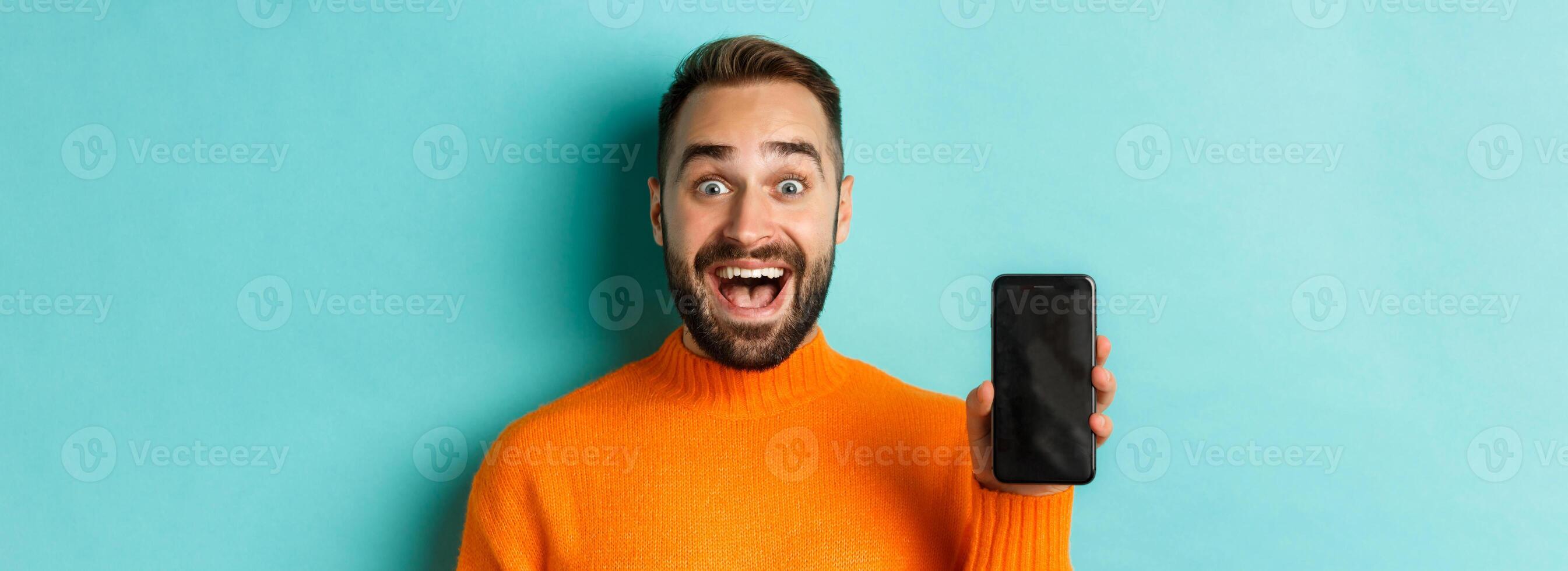 Close-up of handsome bearded guy in orange sweater, showing smartphone screen and smiling, showing promo online, turquoise background photo