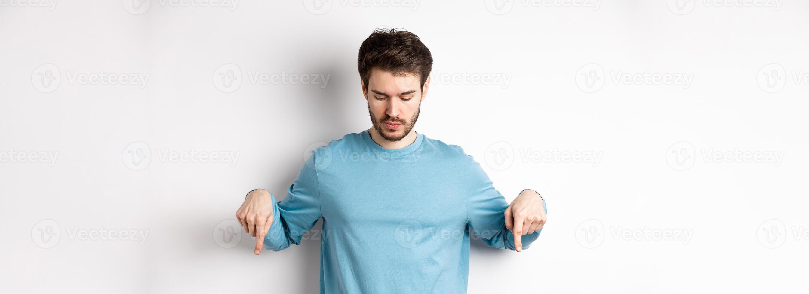 Handsome caucasian man with beard, wearing blue shirt, looking and pointing down at interesting logo banner, standing on white background photo