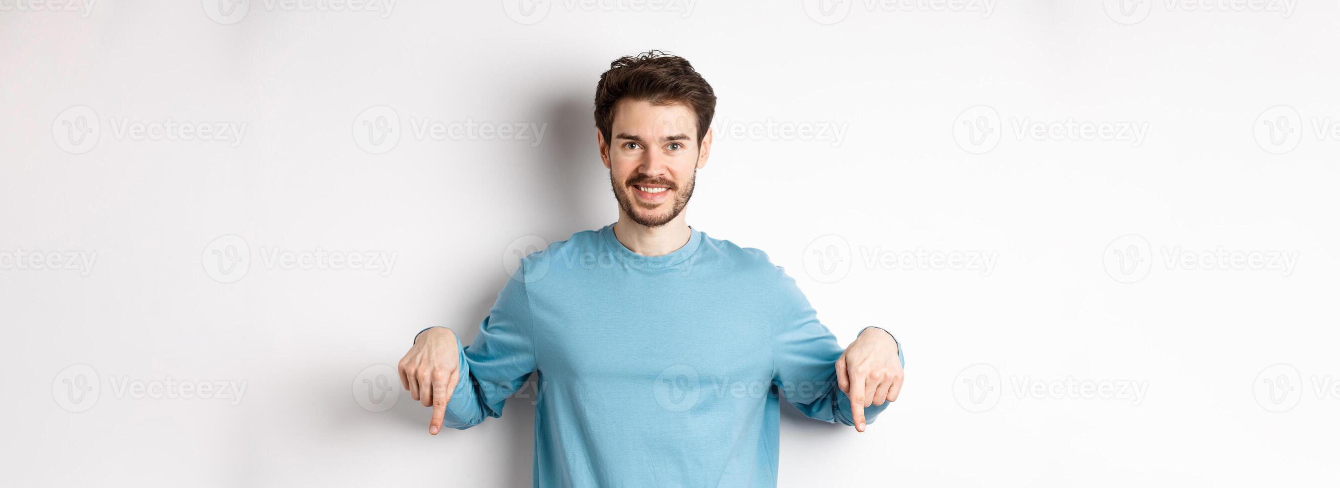 Smiling confident man pointing fingers down, showing promo banner or logo on white background, standing in casual blue sweatshirt photo