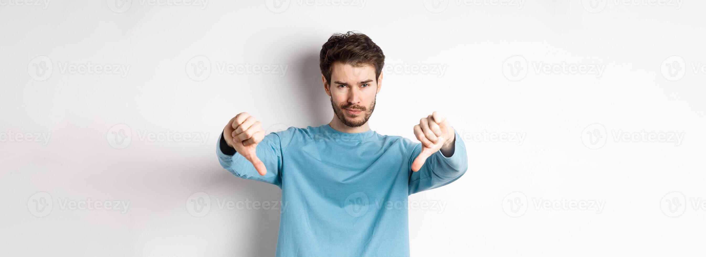 Skeptical handsome guy with beard, showing thumbs down in disapproval, dislike something bad, standing over white background photo