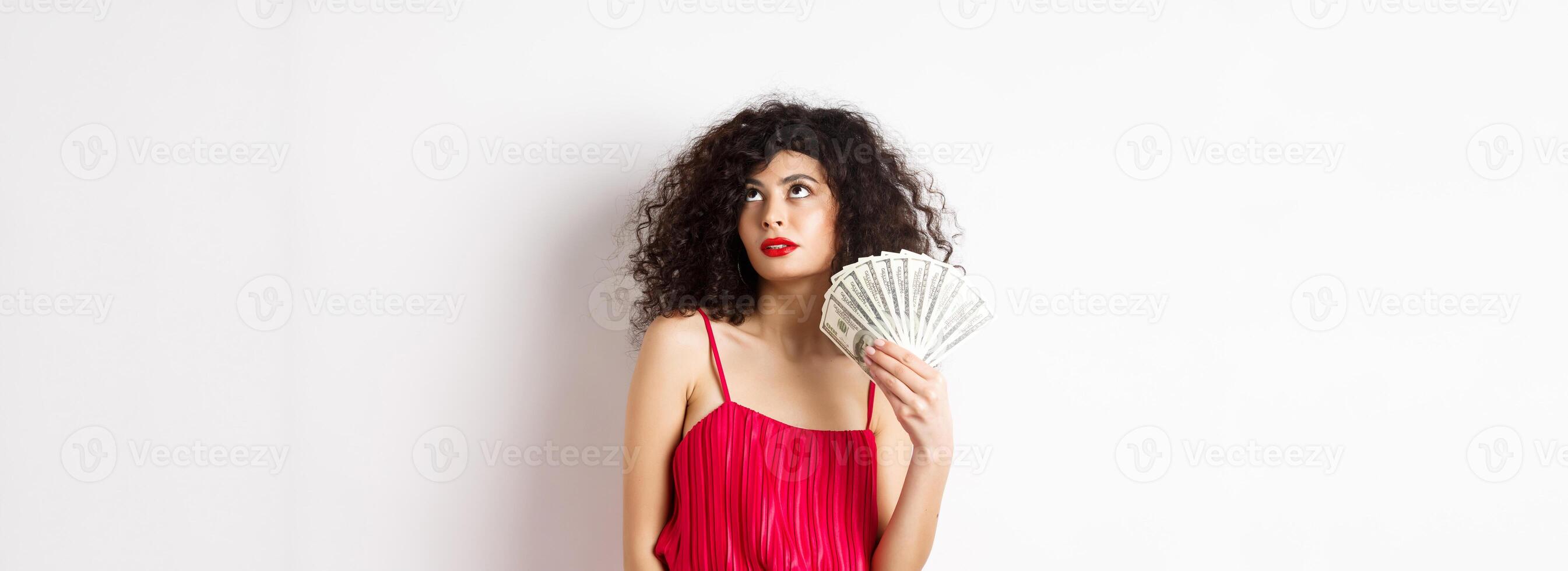 Elegant beautiful woman in red dress, waving with dollars and looking fabulous, standing on white background photo