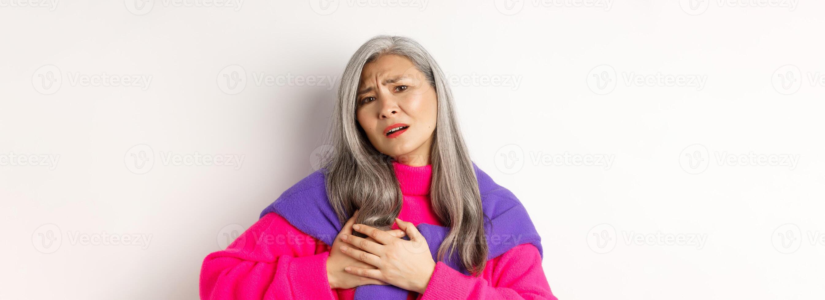 Worried asian mother holding hands on heart and grimacing, looking anxious and having pain in chest, feeling hurt, standing over white background photo