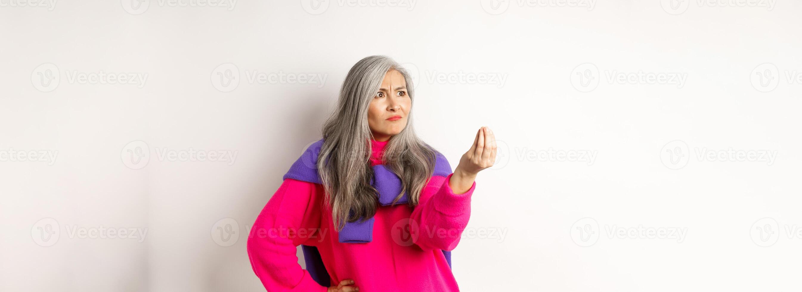 Grumpy asian grandmother complaining, showing italian what do you want gesture, shaking fingers and staring angry left at copy space, standing over white background photo