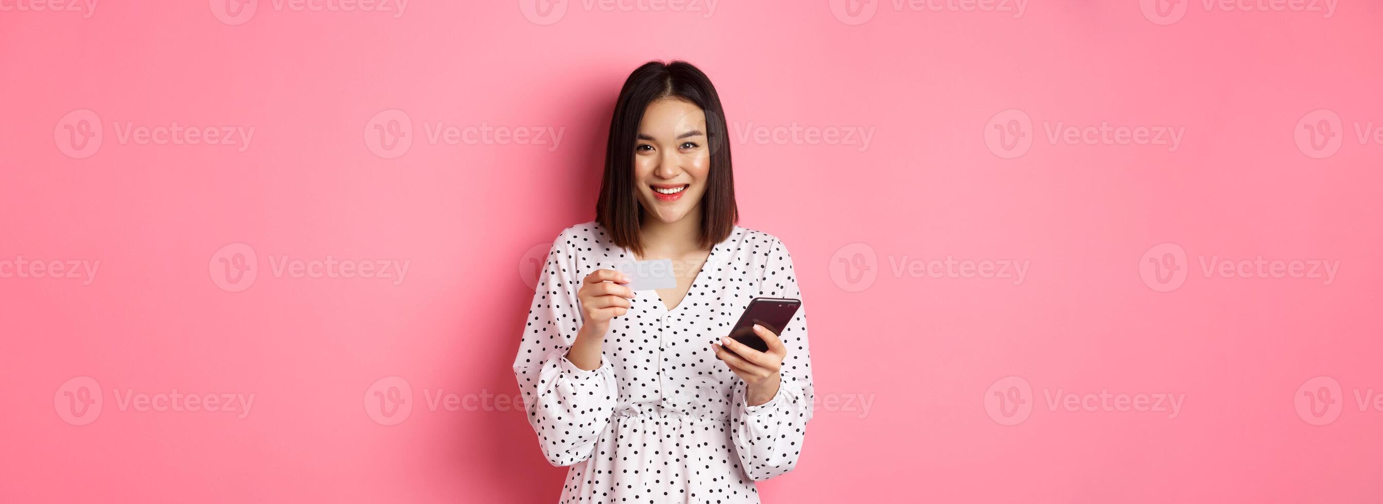 Beautiful asian woman enter credit card info in mobile phone app, shopping online, paying for order on smartphone, standing over pink background photo