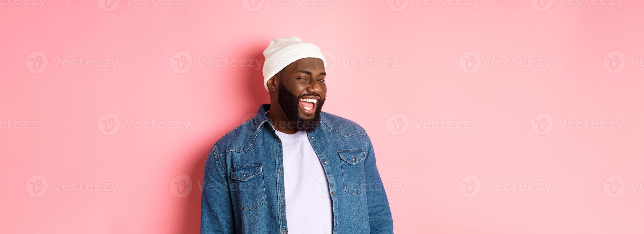 Cheeky african-american man winking at camera and smiling, teasing with offer, standing in hipster clothing against pink background photo