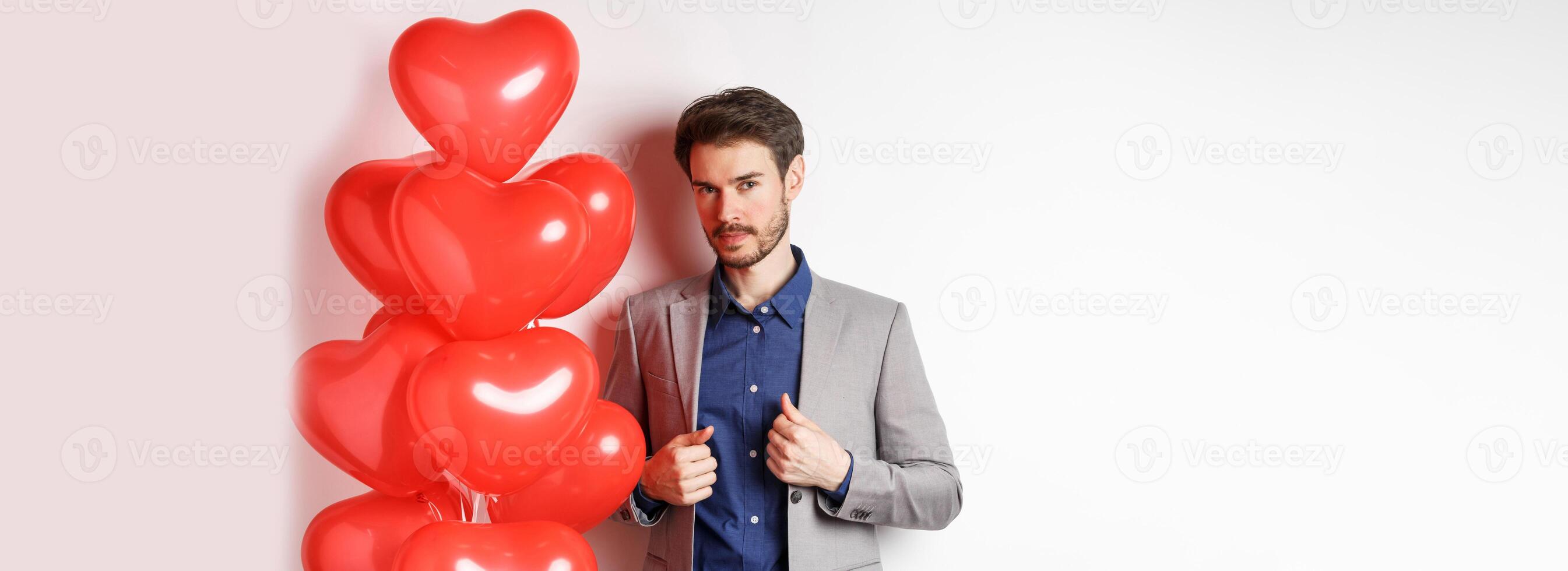 Lovers day. Handsome and confident young man getting dressed for Valentines day, fixing suit and looking at camera, standing near romantic heart balloons, white background photo