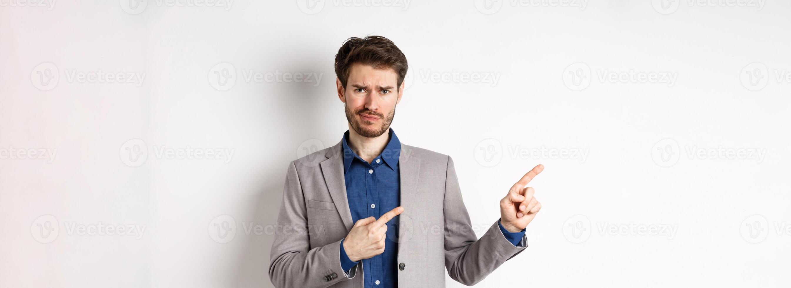 Bad idea. Displeased skeptical guy in business suit smirk and frown, pointing fingers left at upsetting news, looking hesitant at camera, white background photo