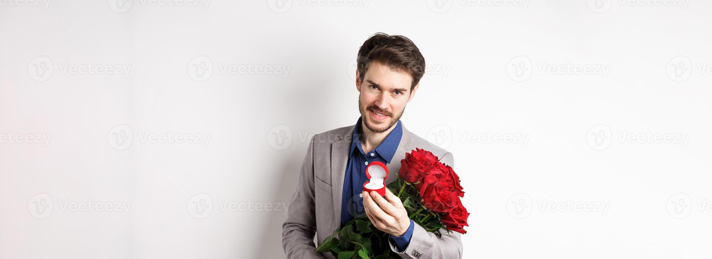 Romantic man with boquet of red roses asking to marry him, holding engagement ring and looking confident at camera, standing in suit over white background photo