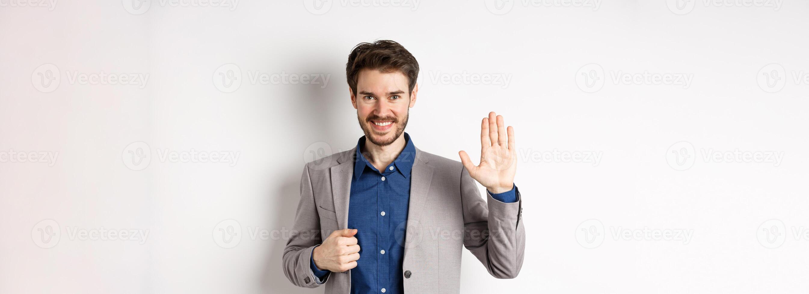 Smiling handsome businessman in suit waving hand, greeting friend with friendly face, say hellp, standing on white background photo