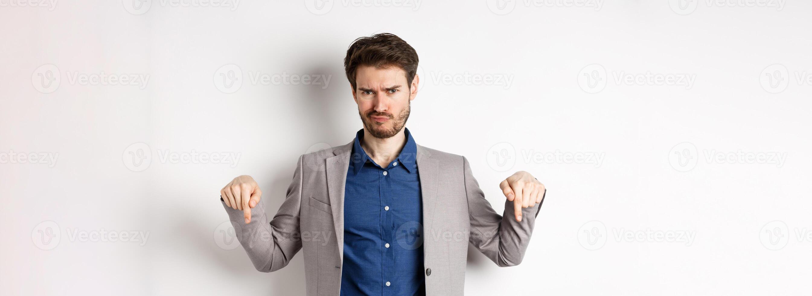 Disappointed bearded male entrepreneur frowning and pointing down with negative emotion, express dislike or bad opinion, standing on white background photo