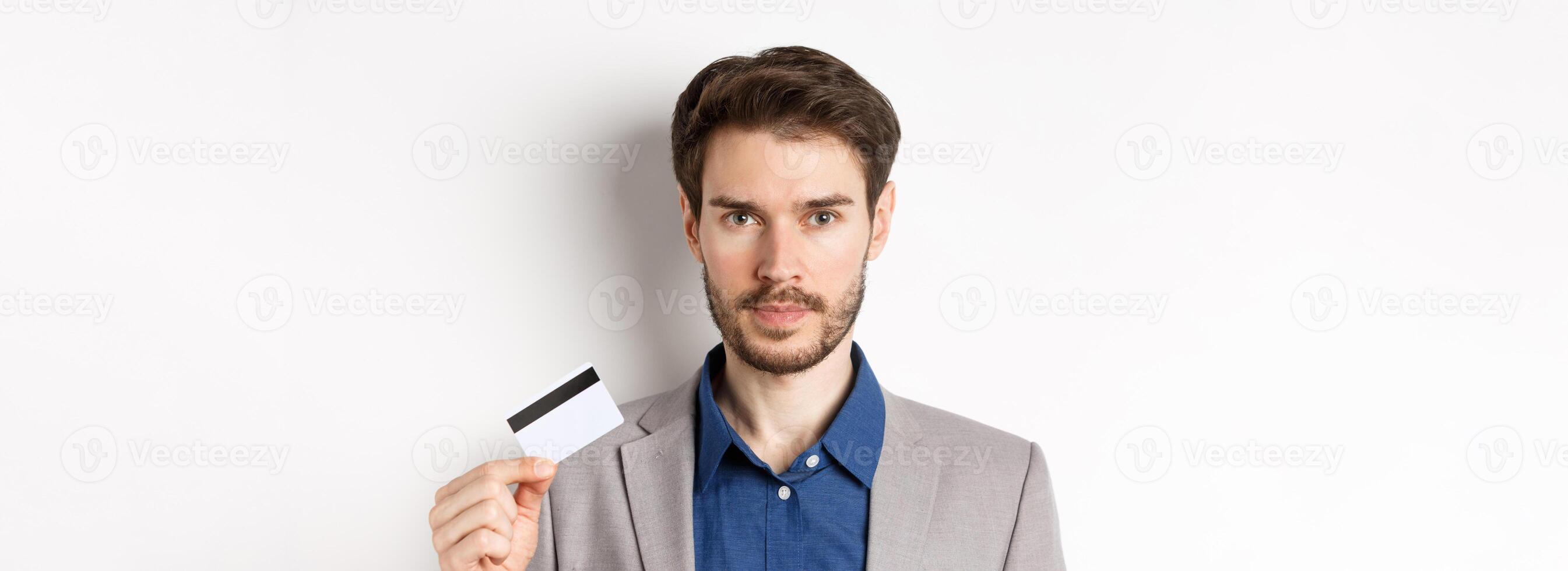 Finance. Close up of handsome bearded businessman in suit showing plastic credit card, standing serious on white background photo
