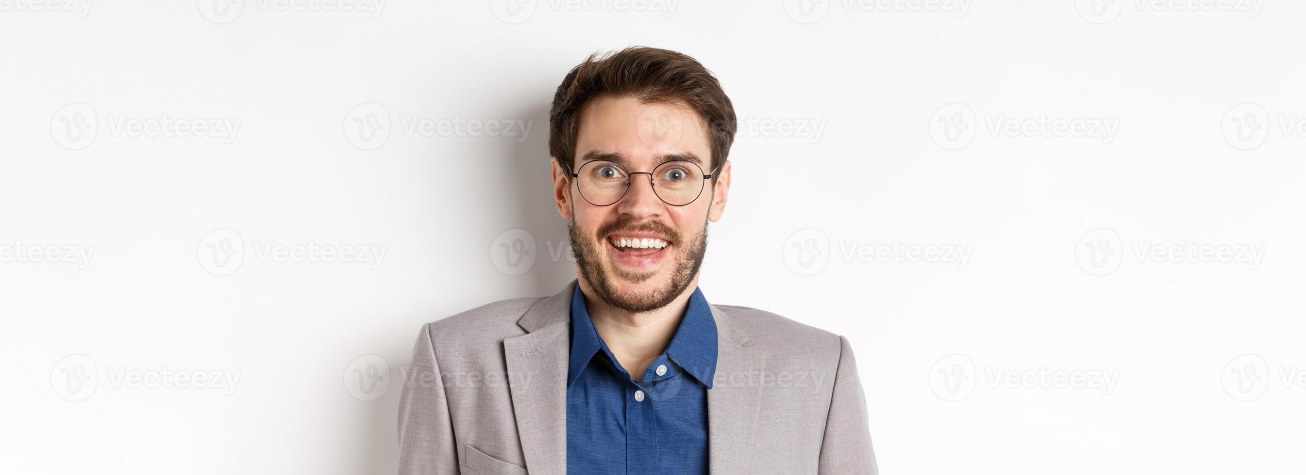 Cheerful european man in glasses and suit smiling excited, looking at camera with amazed happy face, white background photo
