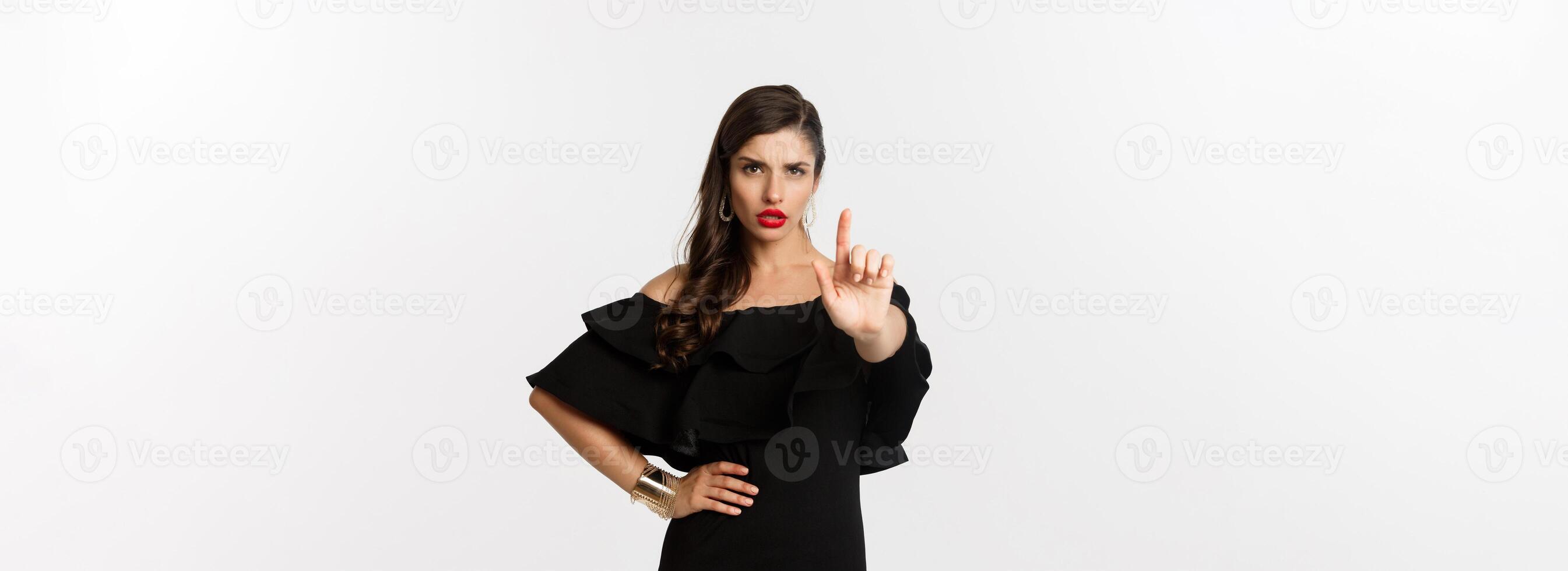 Fashion and beauty. Confident and serious lady in black dress, showing finger in stop gesture, prohibit and disapprove something, standing over white background photo