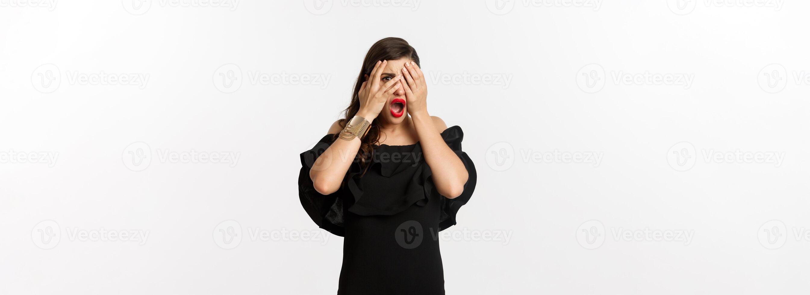 Fashion and beauty. Shocked young woman in black dress covering eyes, peeking through fingers at something embarrassing, cringe, standing over white background photo