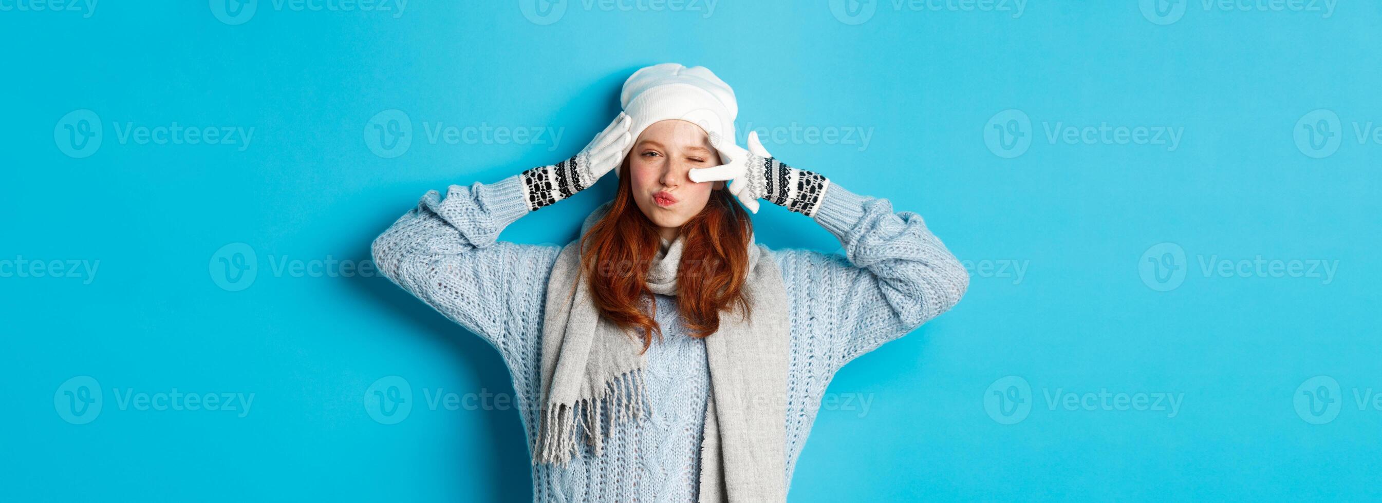 Winter and holidays concept. Happy redhead girl in beanie hat, scarf and gloves showing peace sign and pucker lips sassy, staring at camera, wearing sweater, posing over blue background photo