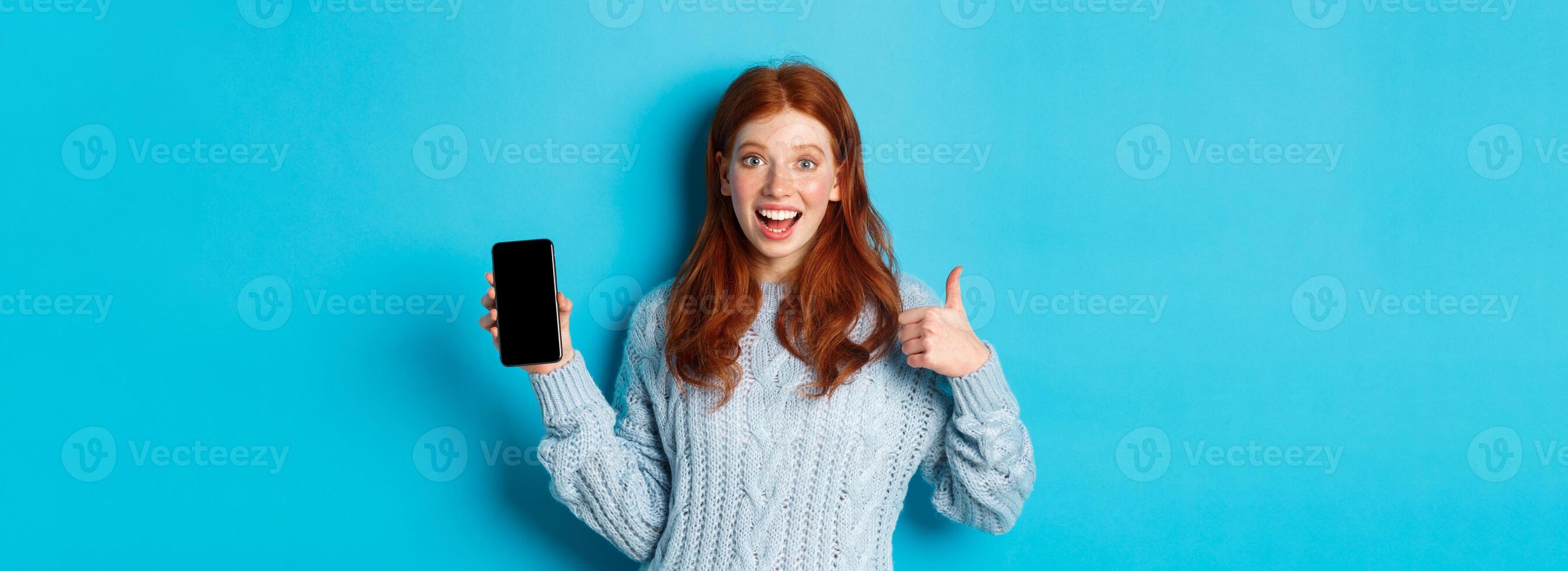 Smiling redhead girl showing smartphone screen, holding phone and demonstrating app, making thumb up in approval, recommending online store, blue background photo