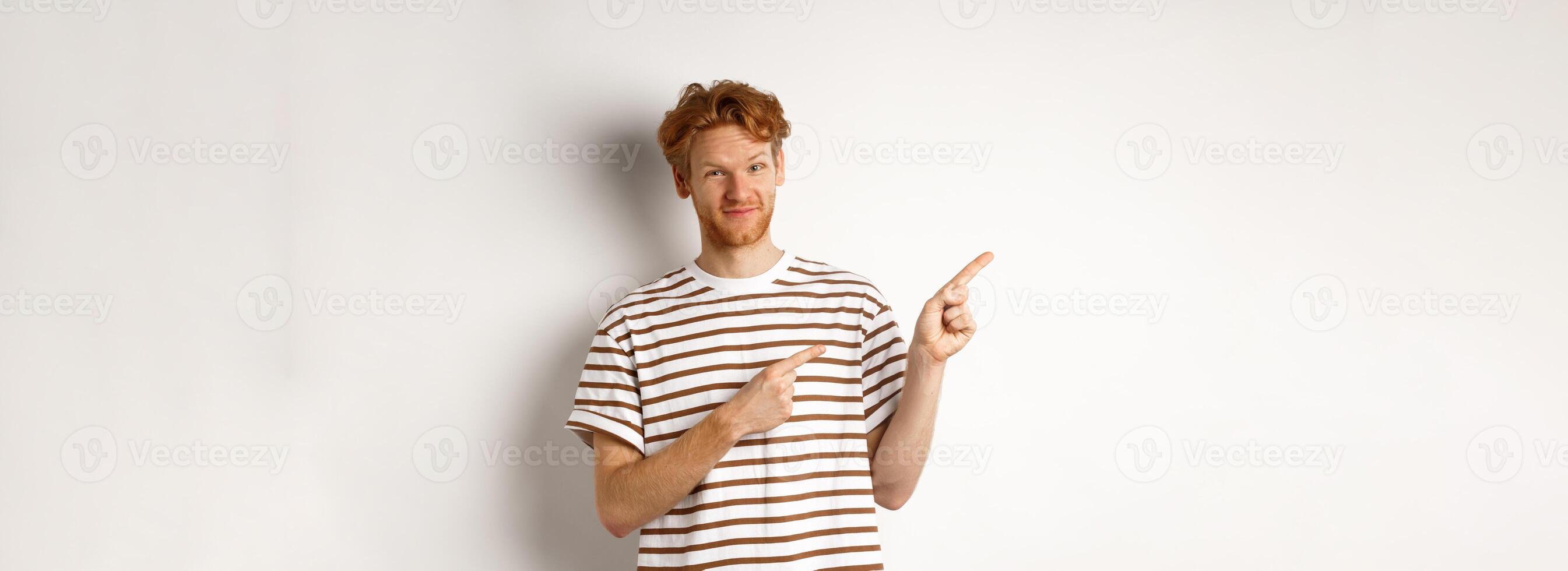 Doubtful redhead guy pointing fingers left at looking skeptical at camera, standing over white background photo