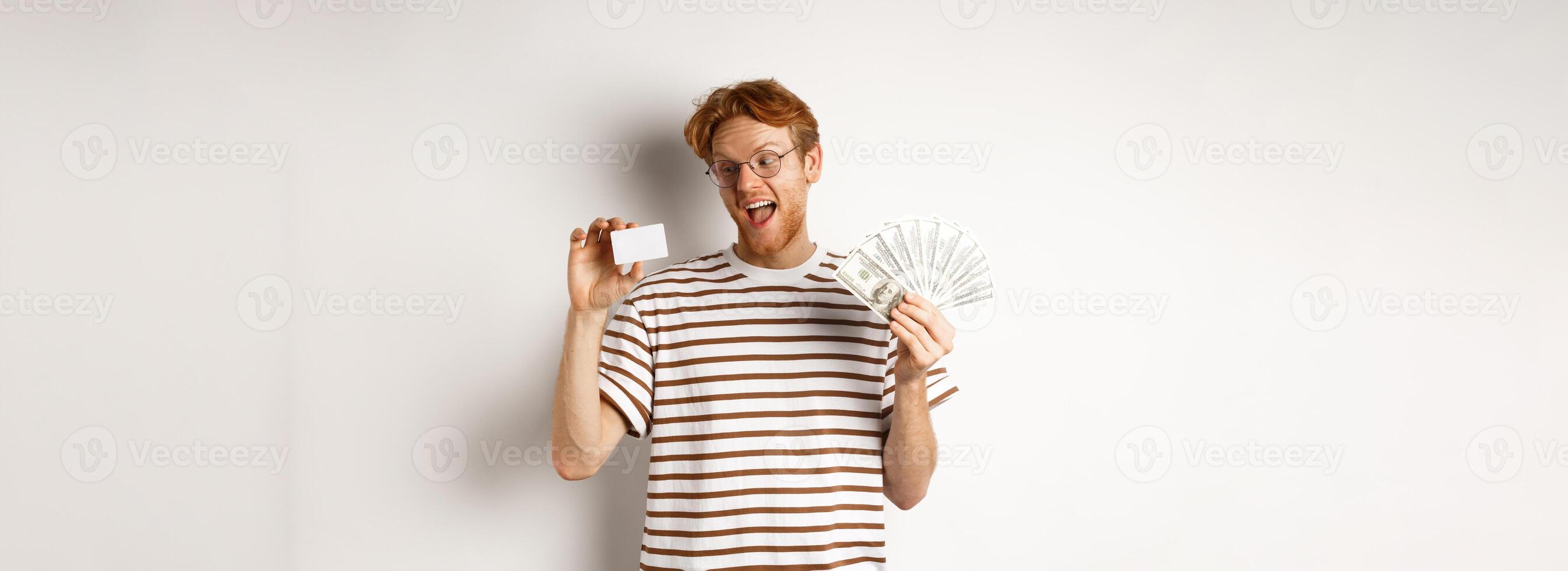 Shopping and finance concept. Young redhead man in glasses choosing plastic credit card, showing dollars and smiling, standing over white background photo