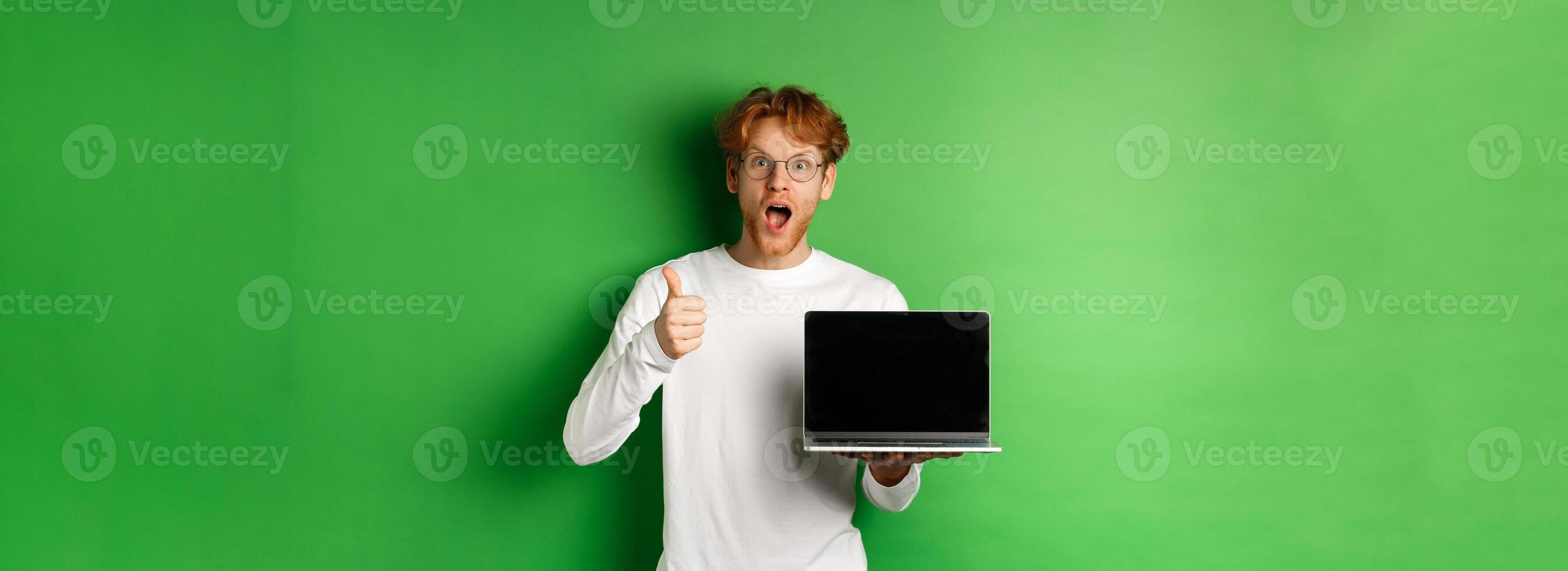 Impressed caucasian man with red hair and beard, showing blank laptop screen and thumb-up, looking amazed at camera, praising something online, green background photo