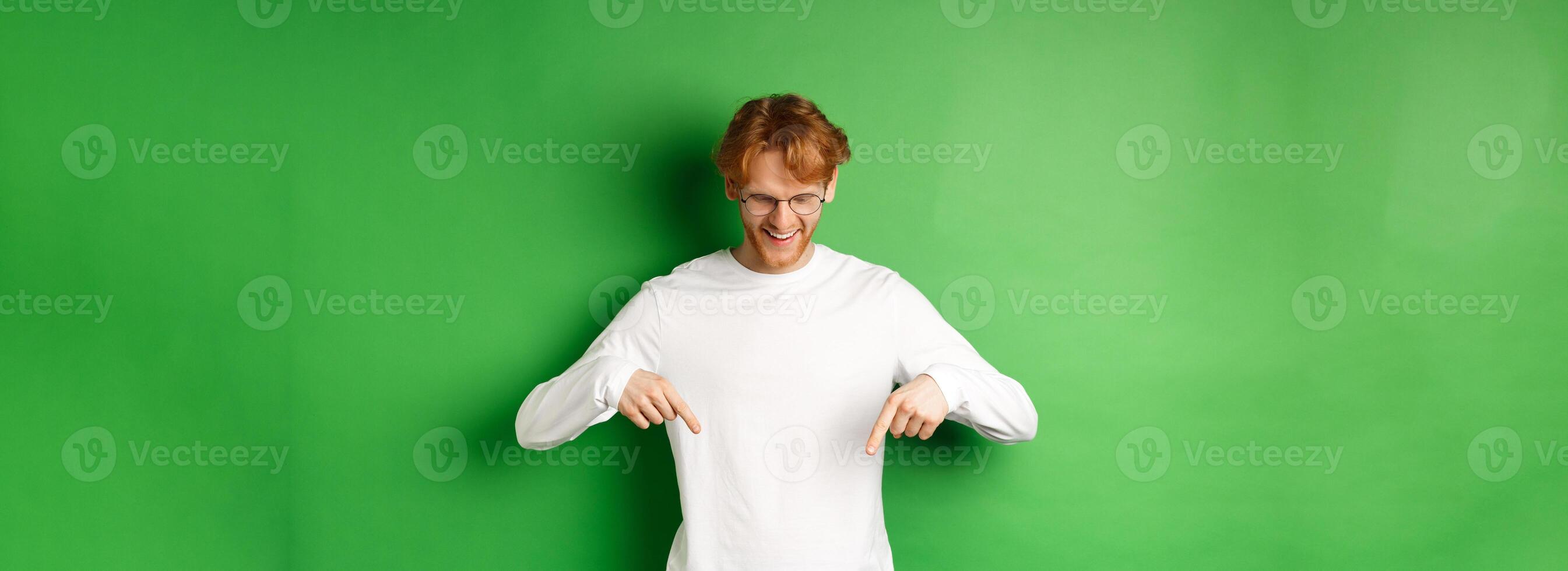 Handsome redhead man in glasses and white long-sleeve checking out advertisement, pointing fingers and looking down with pleased smile, standing over green background photo