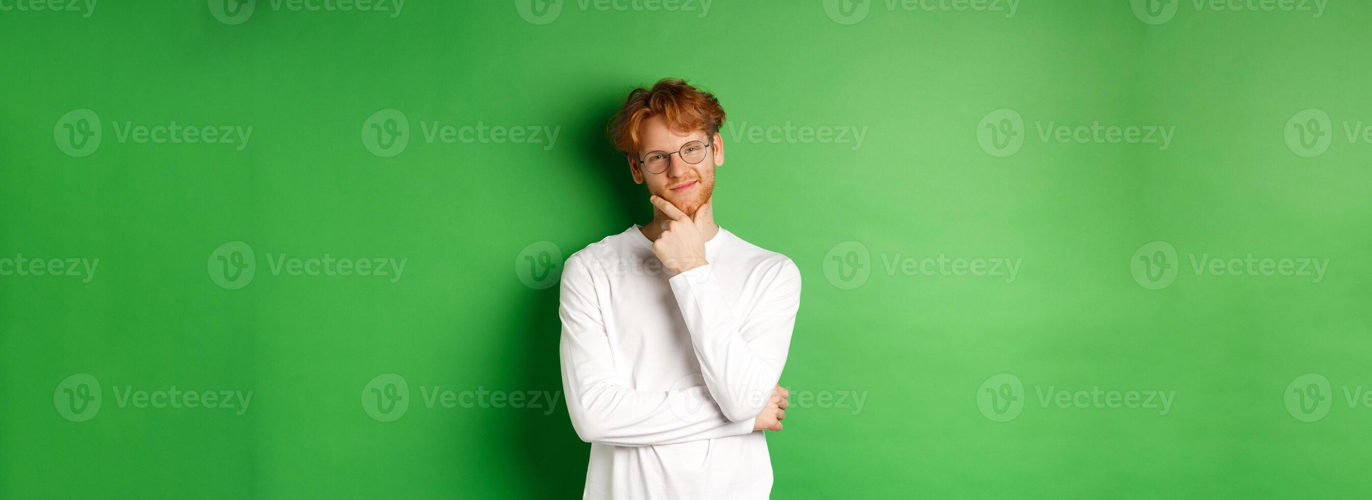 Intrigued young man with red hair, wearing glasses, looking at camera pleased and thoughtful, having an idea, standing over green background photo