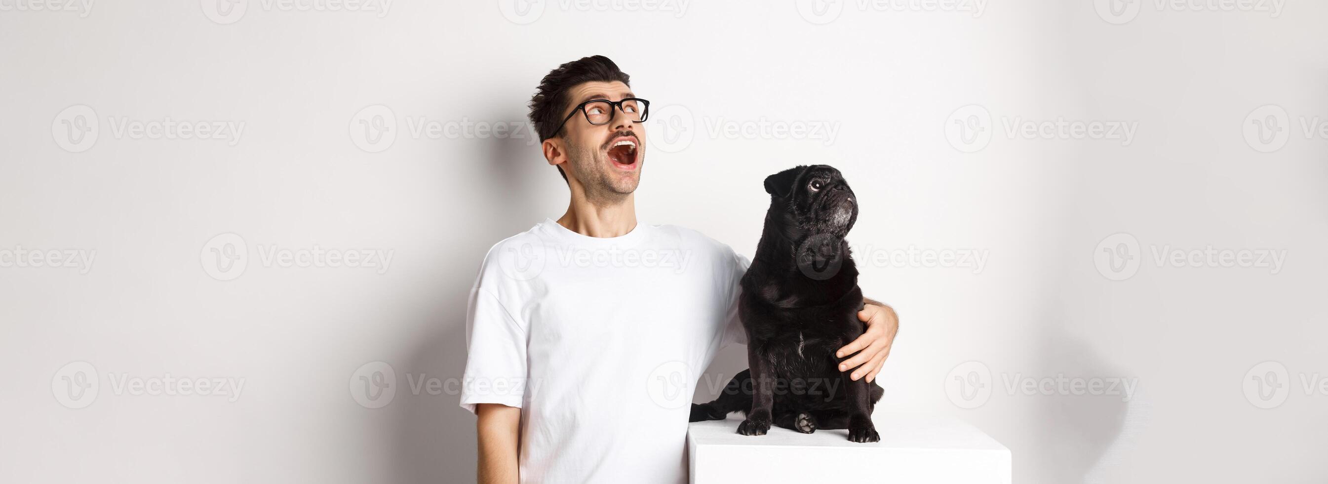 Amazed young man in glasses hugging his dog, pet owner and pug staring at upper left corner promo offer, standing over white background photo