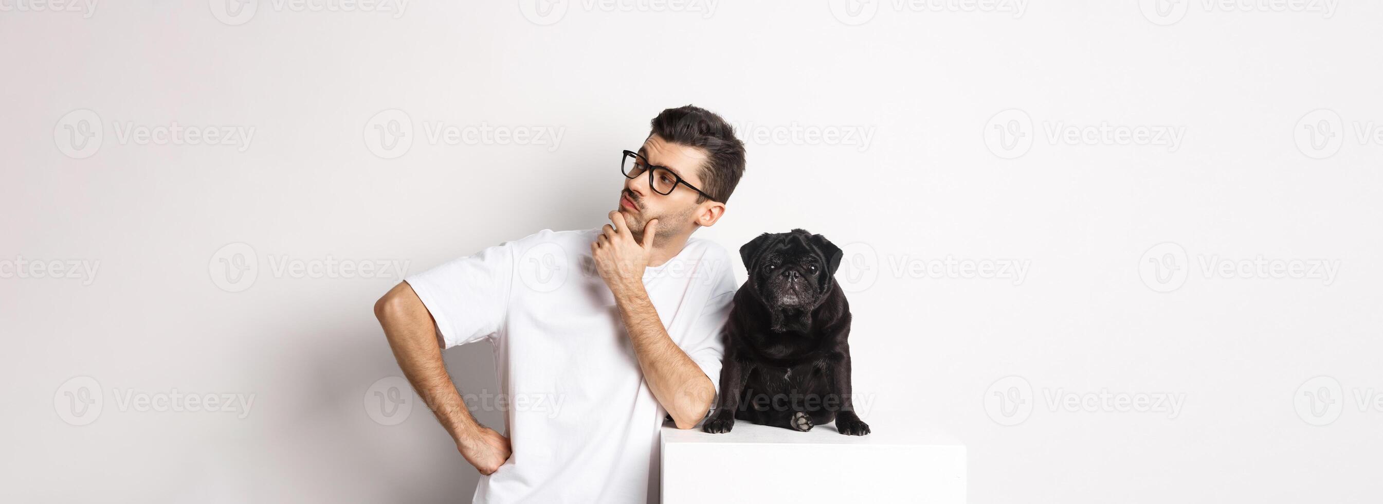 Image of handsome young man, pet owner standing with his dog and looking right at logo, posing with pug against white background photo