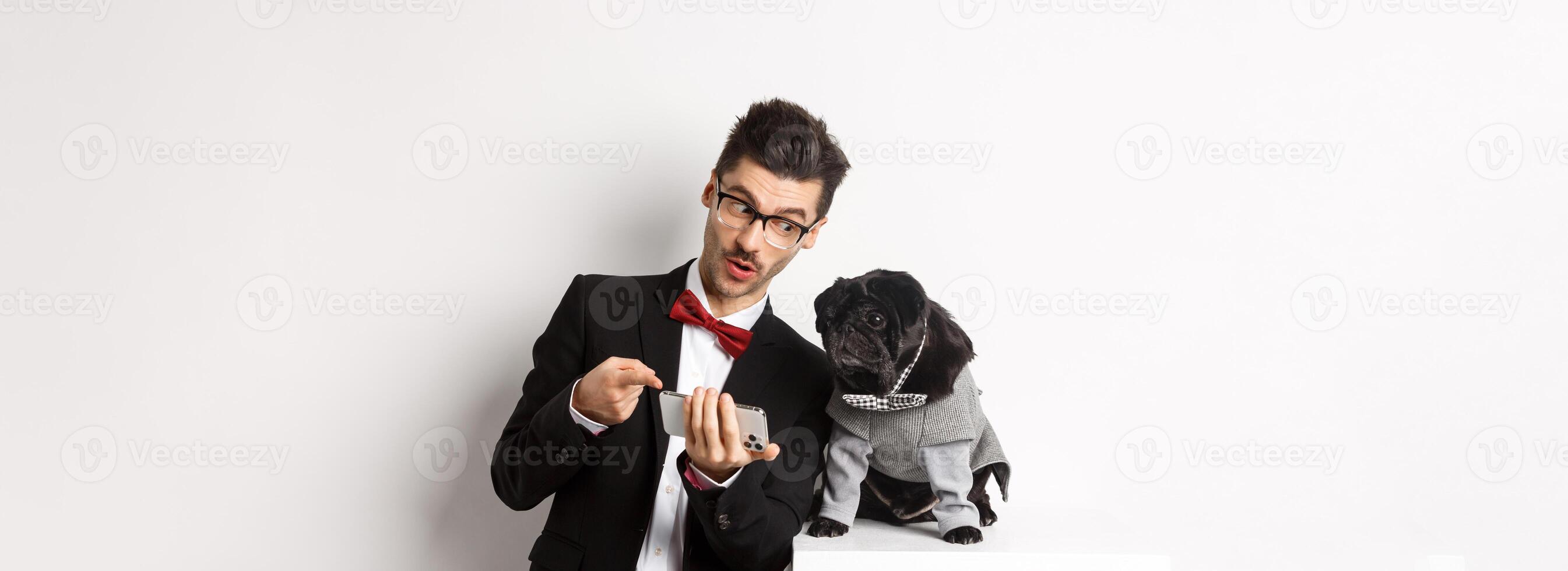 Handsome young man showing something on mobile phone to his dog. Owner shopping online with pet, standing in costumes over white background photo
