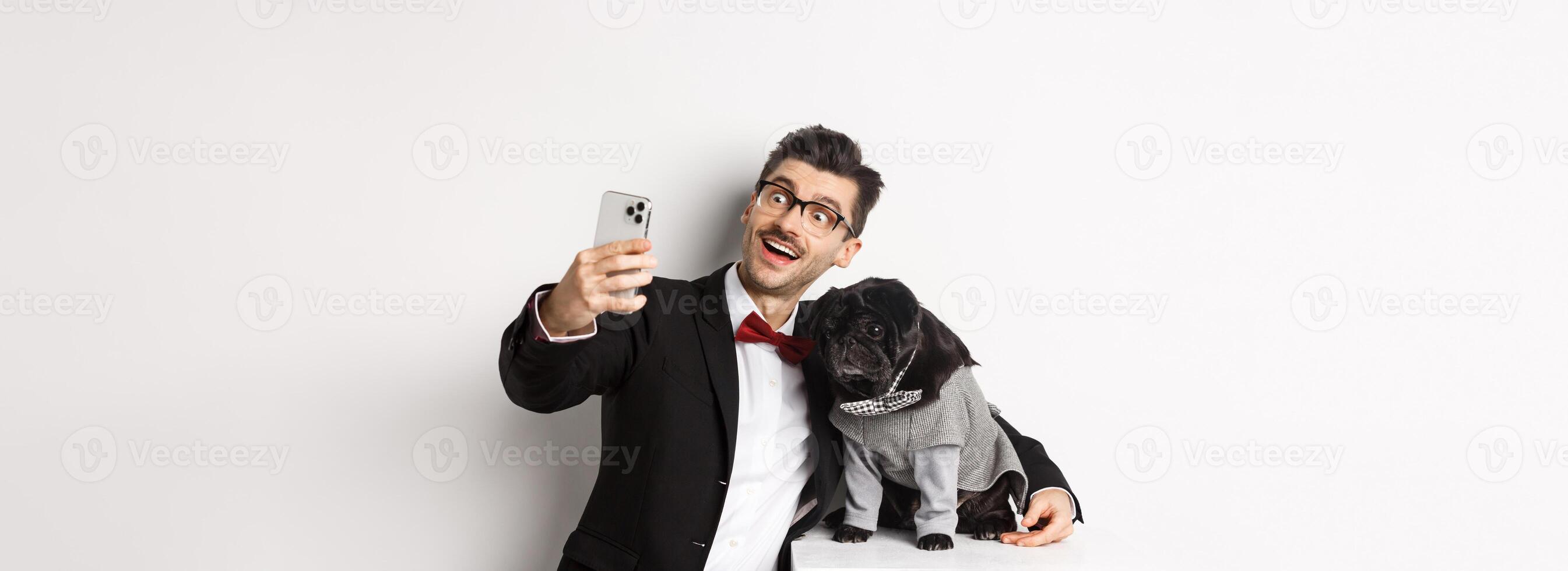 Cheerful dog owner in suit celebrating Christmas with dog, taking selfie on smartphone and hugging cute black pug in costume, white background photo