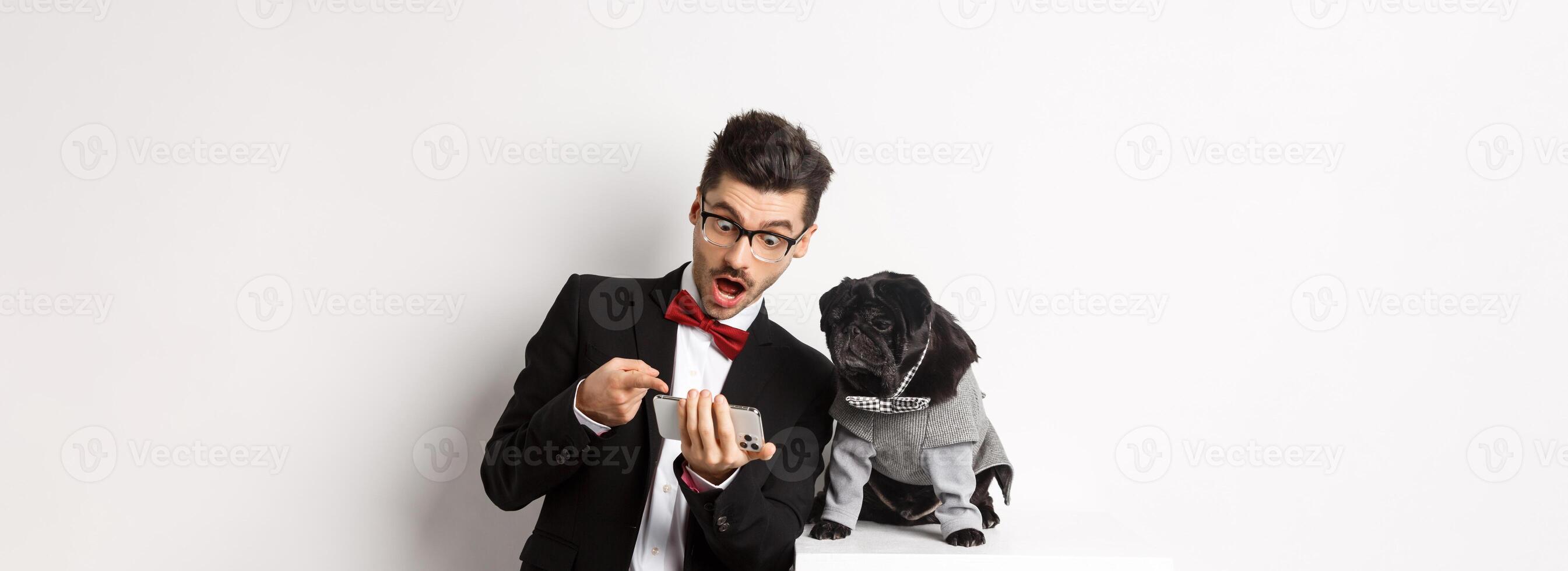 Amazed man showing to dog, pointing at mobile phone and look excited while pug sitting unbothered in costume, white background photo
