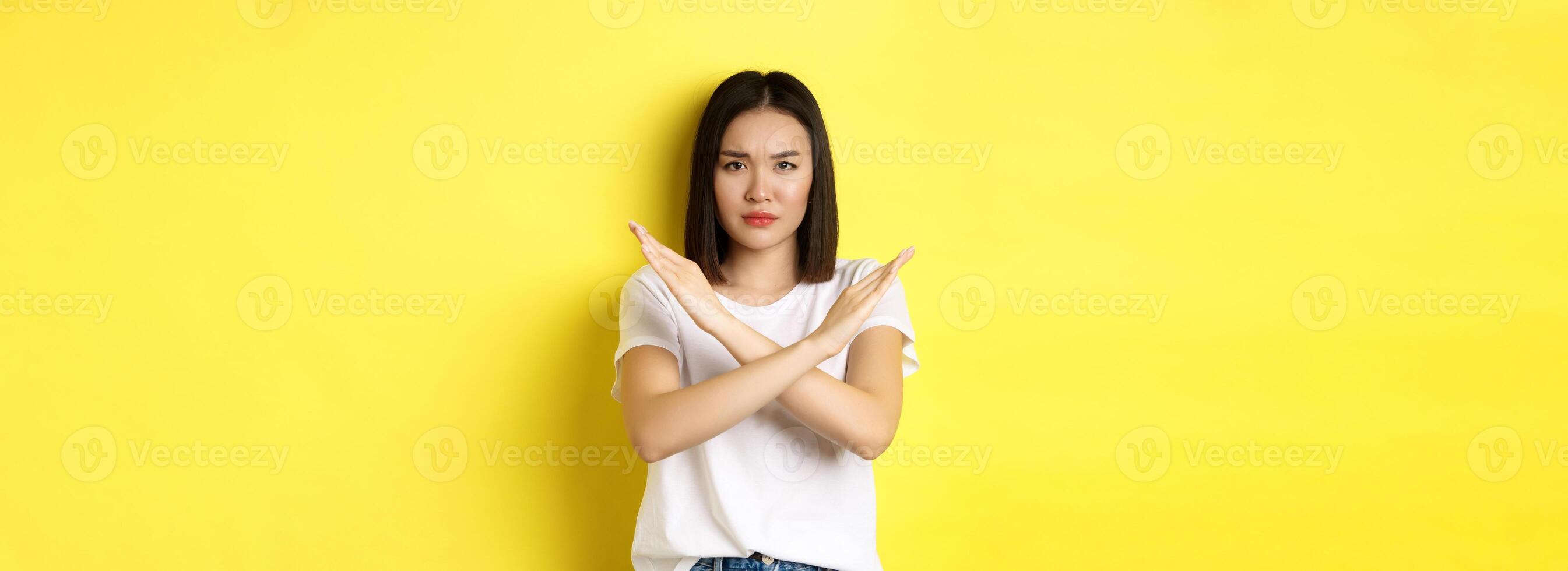 Serious and assertive korean woman showing cross stop gesture, frowning and telling no, prohibit action, disapprove something bad, standing over yellow background photo