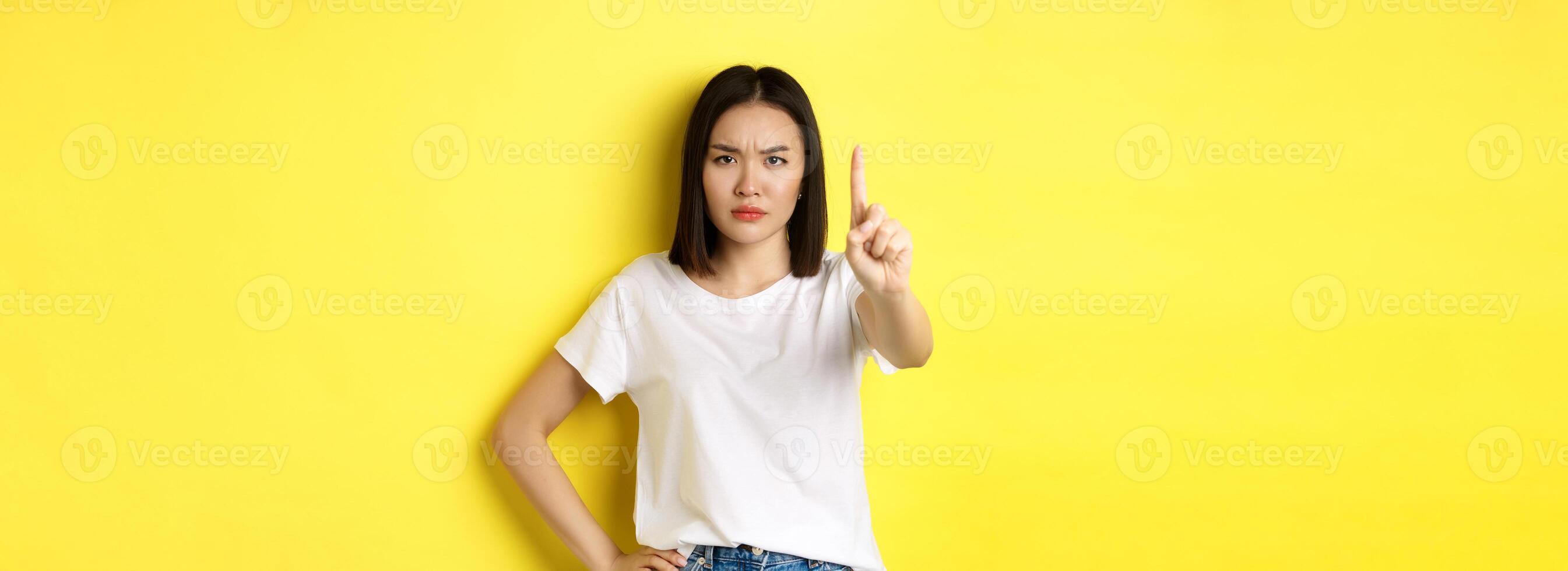 Confident and serious woman tell no, showing extended finger to stop and prohibit something bad, frowning and looking at camera self-assured, standing over yellow background photo