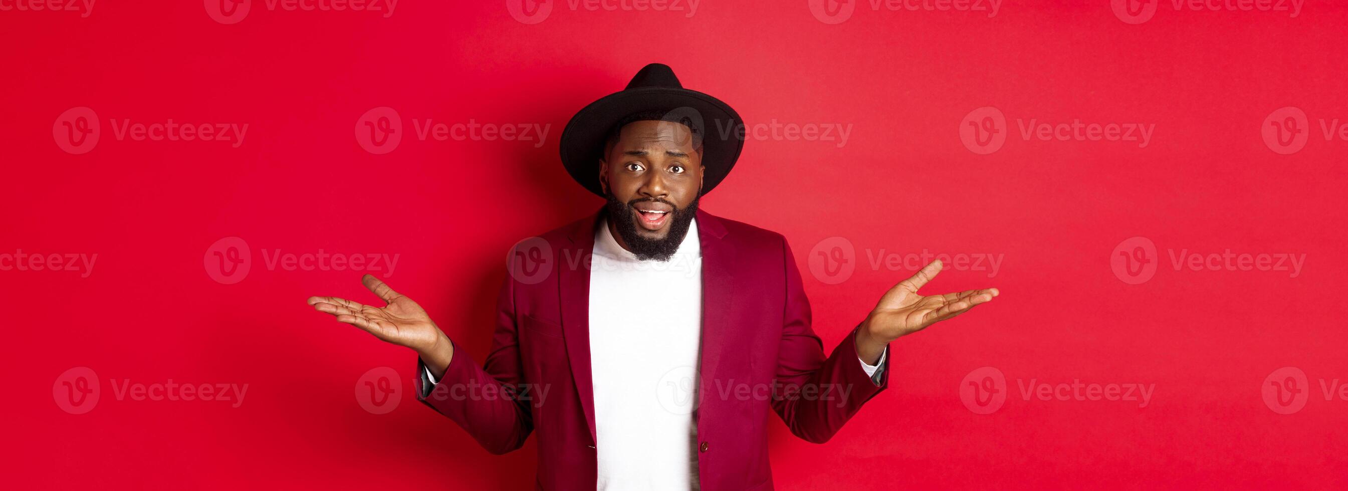 Image of confused Black man asking question, spread hands sideways and staring camera clueless, standing against red background photo
