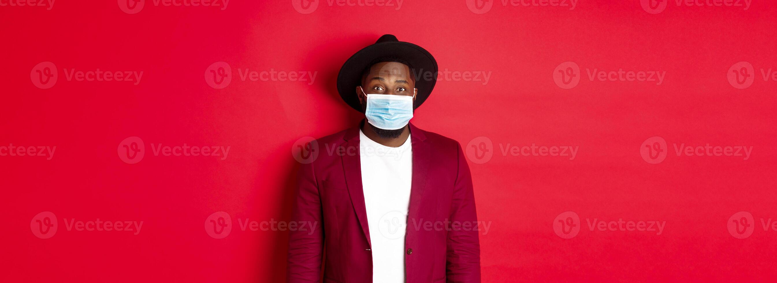 Covid-19 and fashion concept. Handsome african american man in stylish blazer and hat, wearing medical mask from coronavirus, standing over red background photo