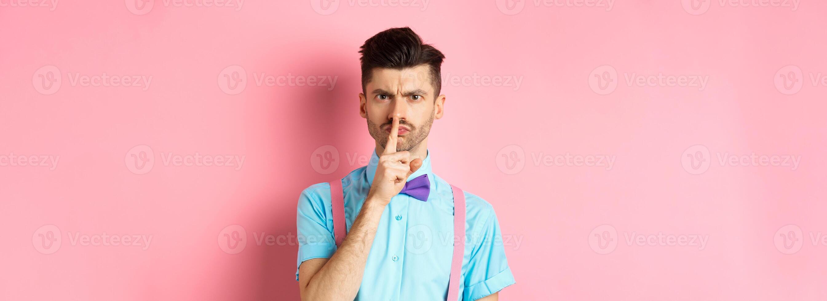 Angry and grumpy man in funny bow-tie shushing at camera, frowning and demand silence, tell to be quiet, scolding someone loud, standing on pink background photo