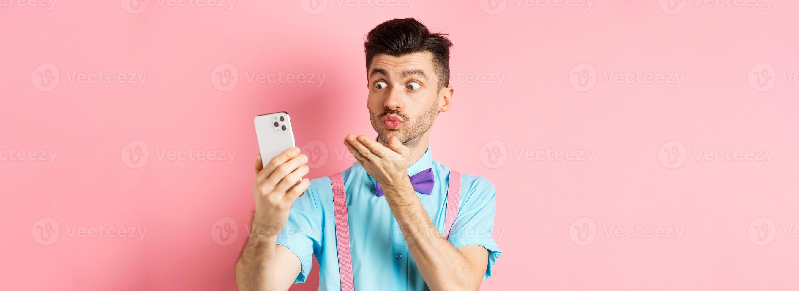 Technology concept. Funny man sending air kiss on chat, blowing mwah at smartphone camera, standing on pink background photo