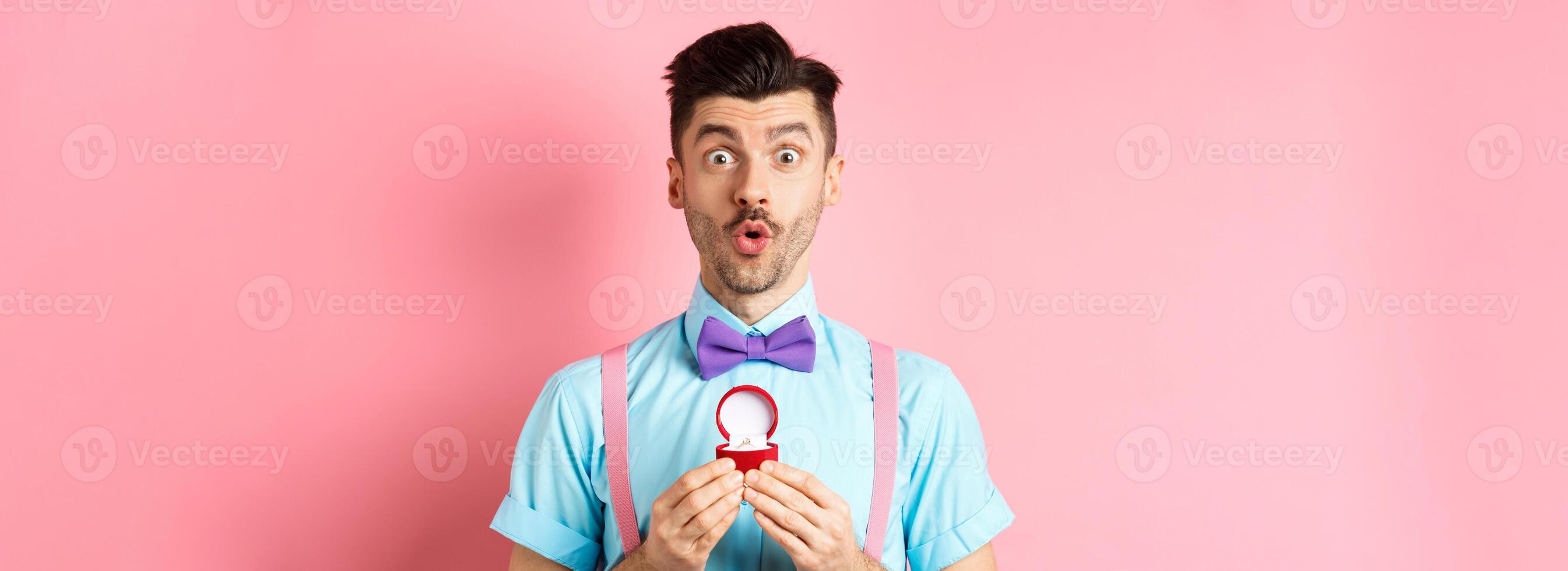 Valentines day. Funny man with moustache and bow-tie, looking excited and showing engagement ring, asking girlfriend to marry him, standing over pink background photo