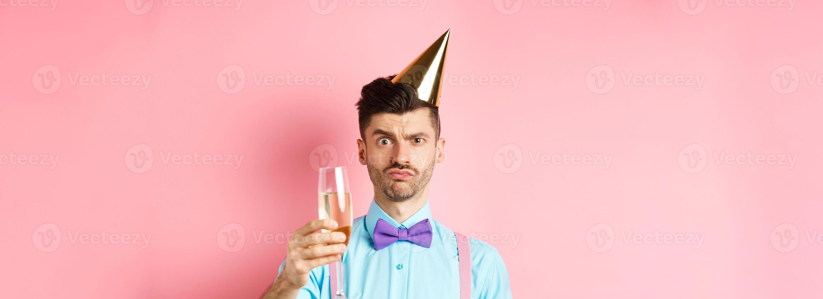 Holidays and celebration concept. Troubled young man in party hat, frowning with doubtful face, raising glass of champagne perplexed, standing on pink background photo
