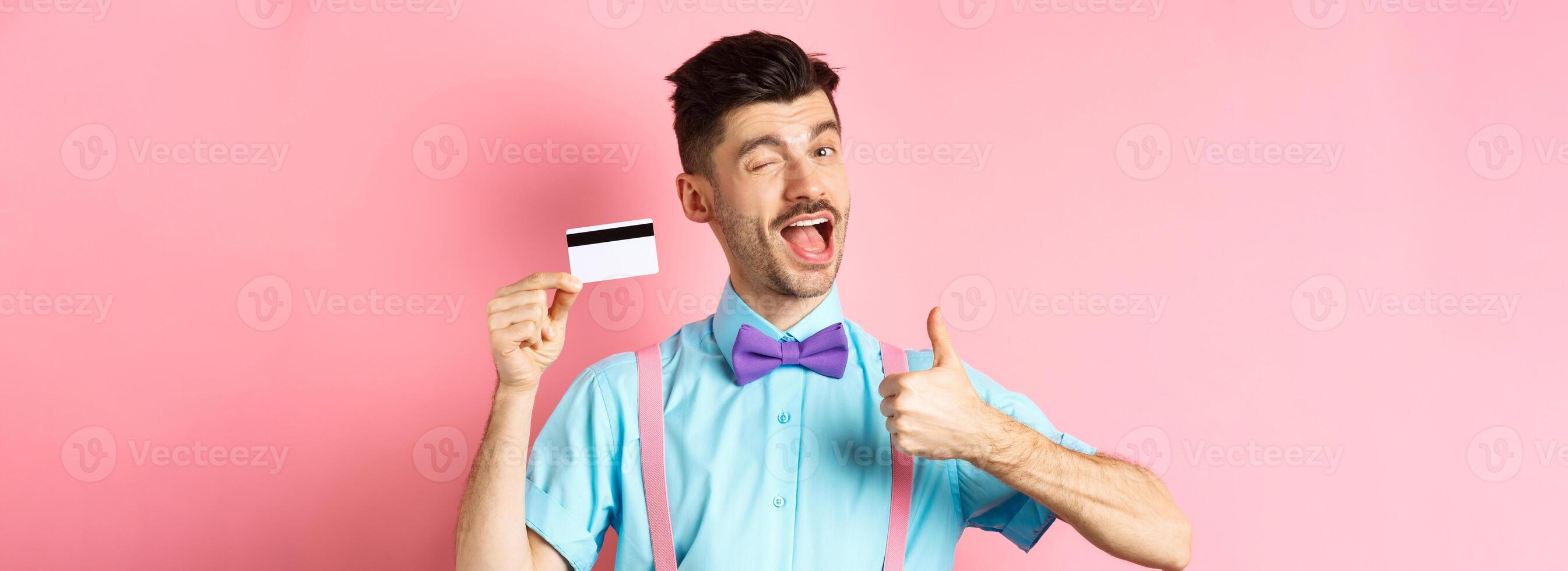 Shopping concept. Festive guy in bow-tie winking at camera, showing thumbs up with plastic credit card, recommending good shop offer, promote bank, standing on pink background photo