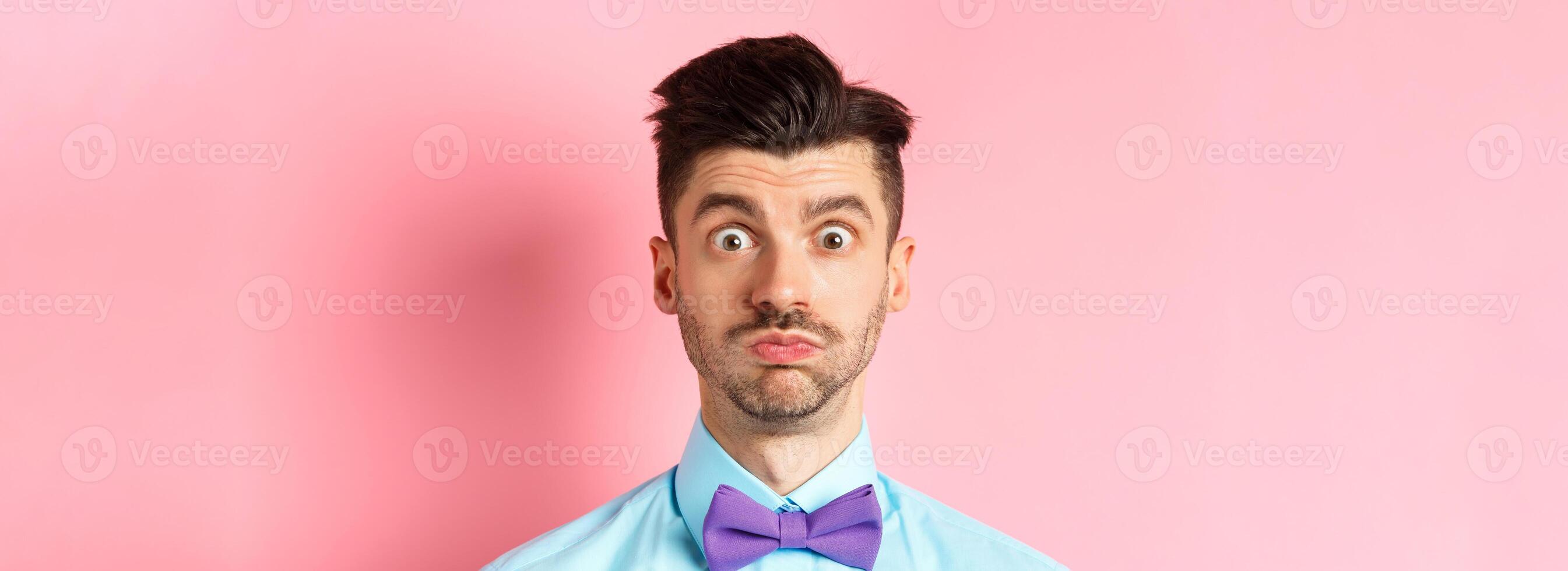 Close-up of confused caucasian guy holding breath, pouting and staring shocked at camera, wearing festive clothes and bow-tie, pink background photo